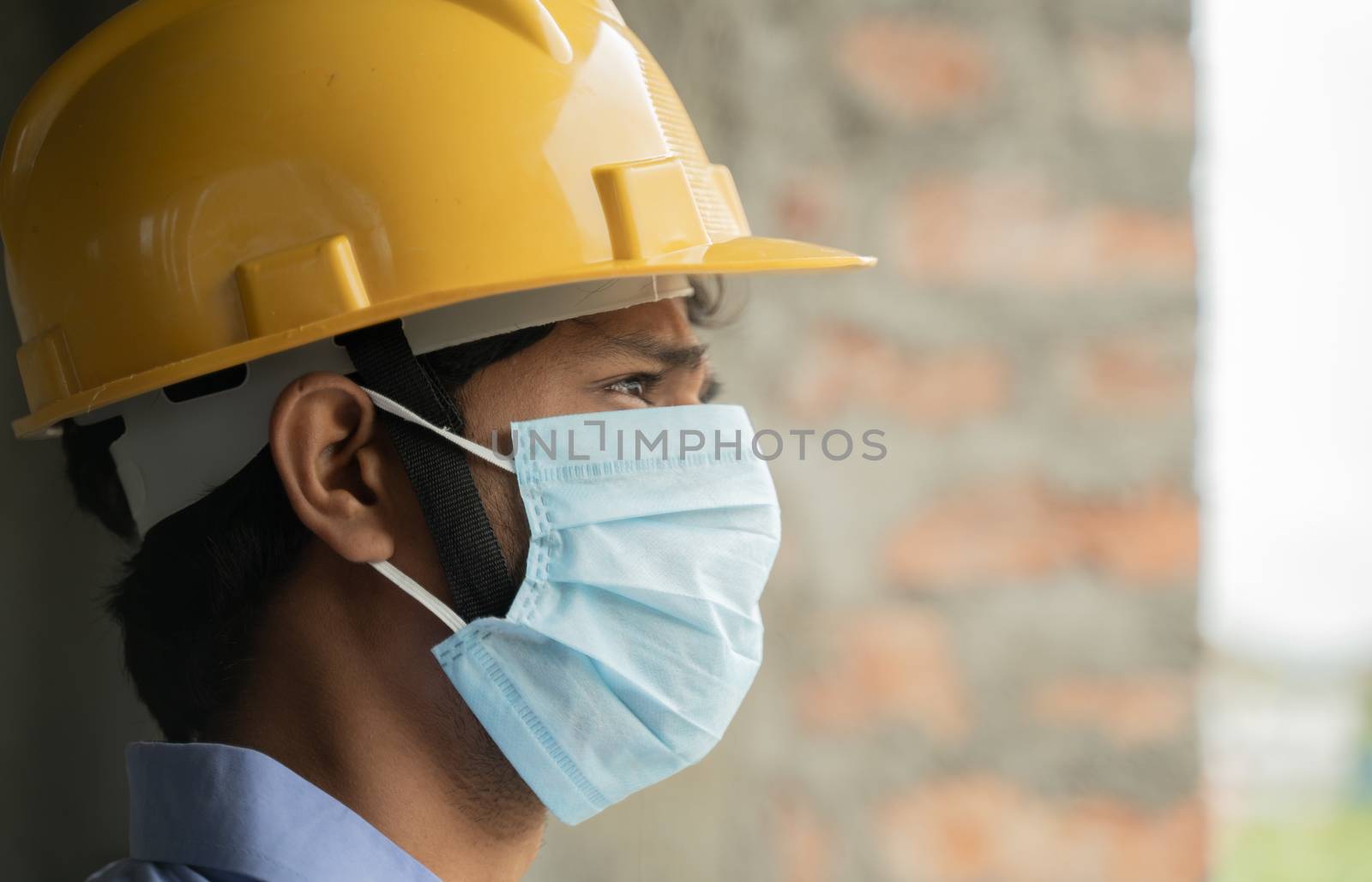 Head shot side view of construction worker seeing outside from construction building window in sad - Industrial worker in a hardhat with medical mask due to coronavirus or covid-19 crisis. by lakshmiprasad.maski@gmai.com