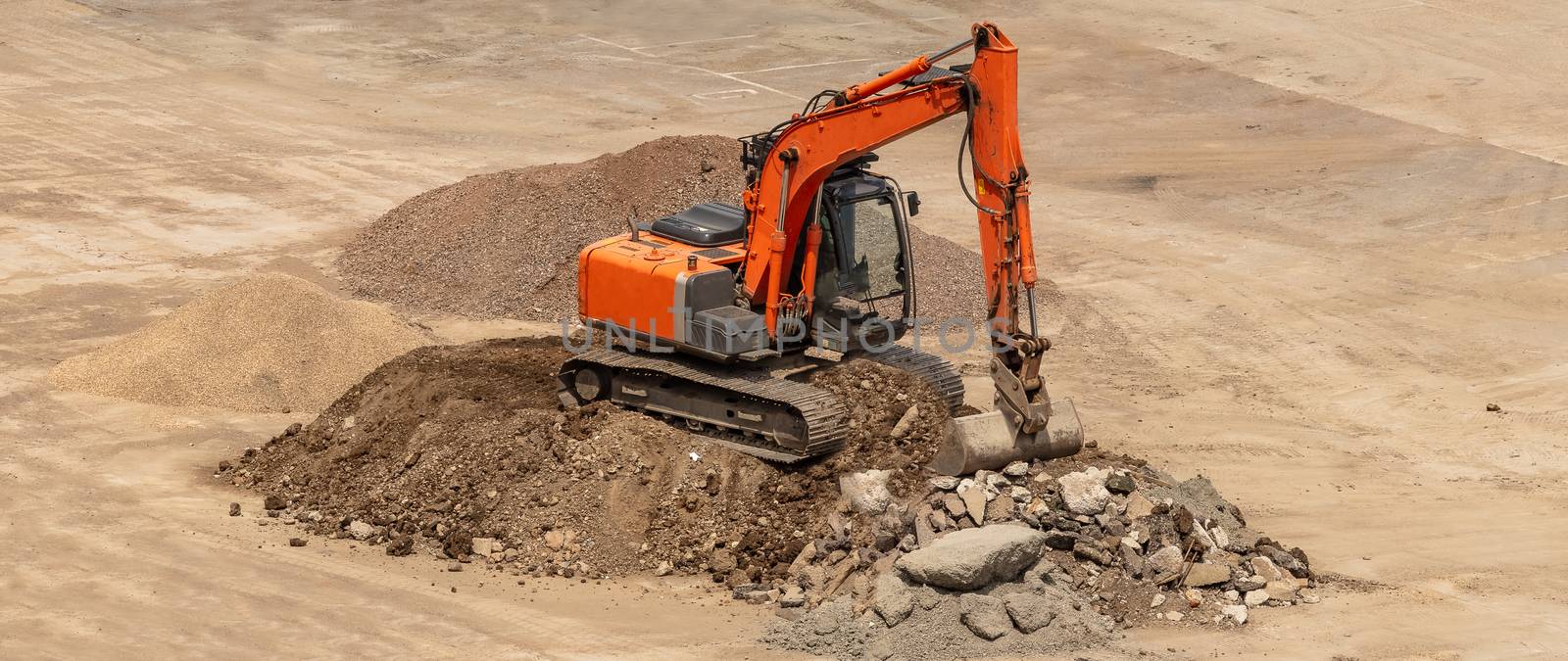 Old orange excavator parked on top of a pile of soil and ready to dig. Rocks lying around. Construction site. Aerial angled view.