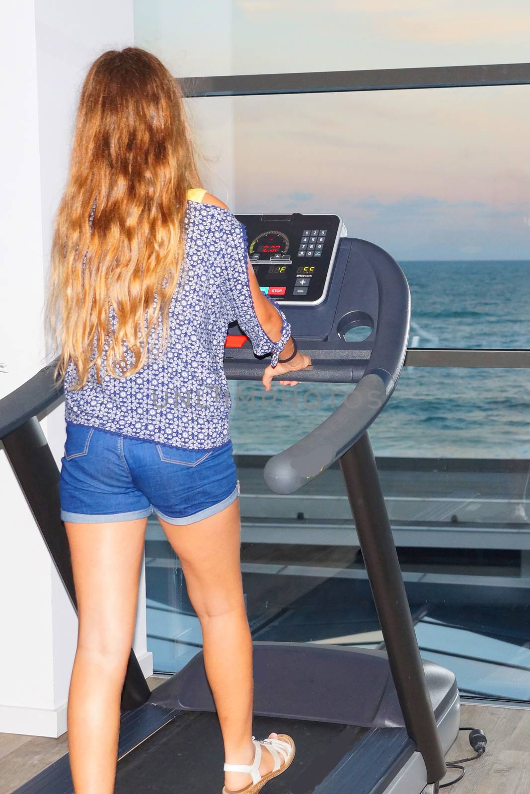 teenage girl on a treadmill in the gym in front of the window with a sea view, back view by Annado