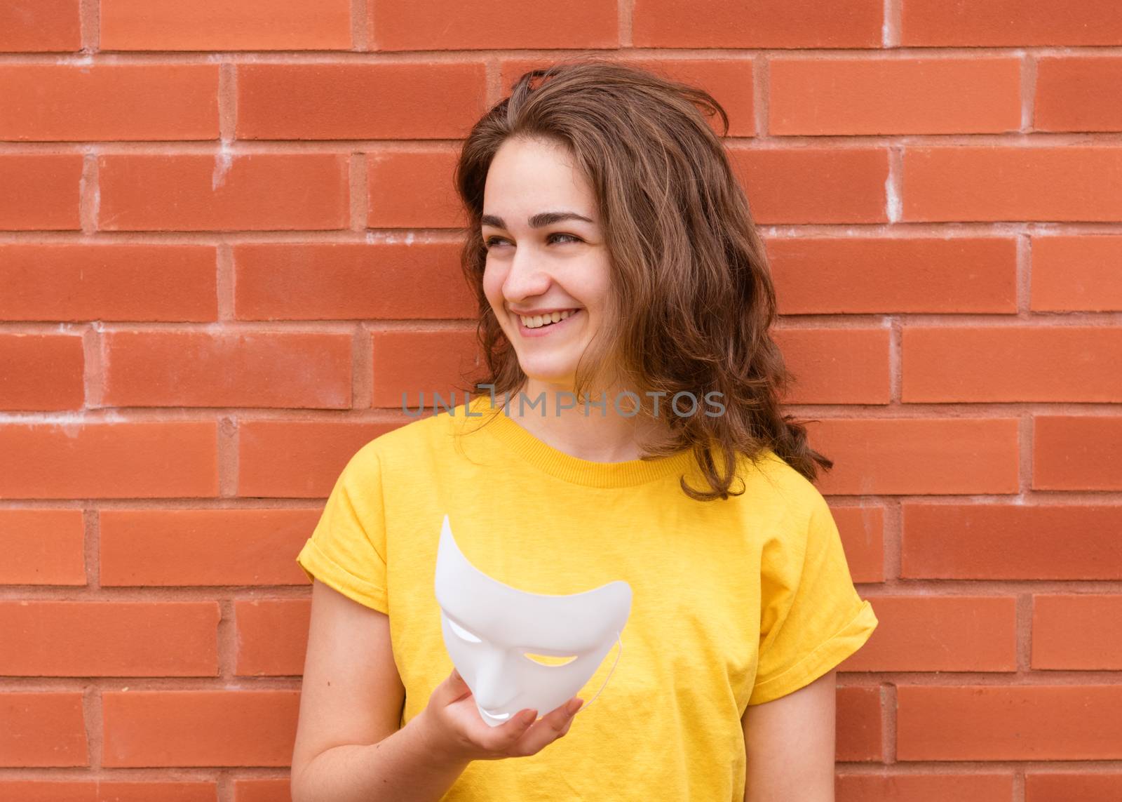 Young woman in yellow shirt putting on a mask against brick wall