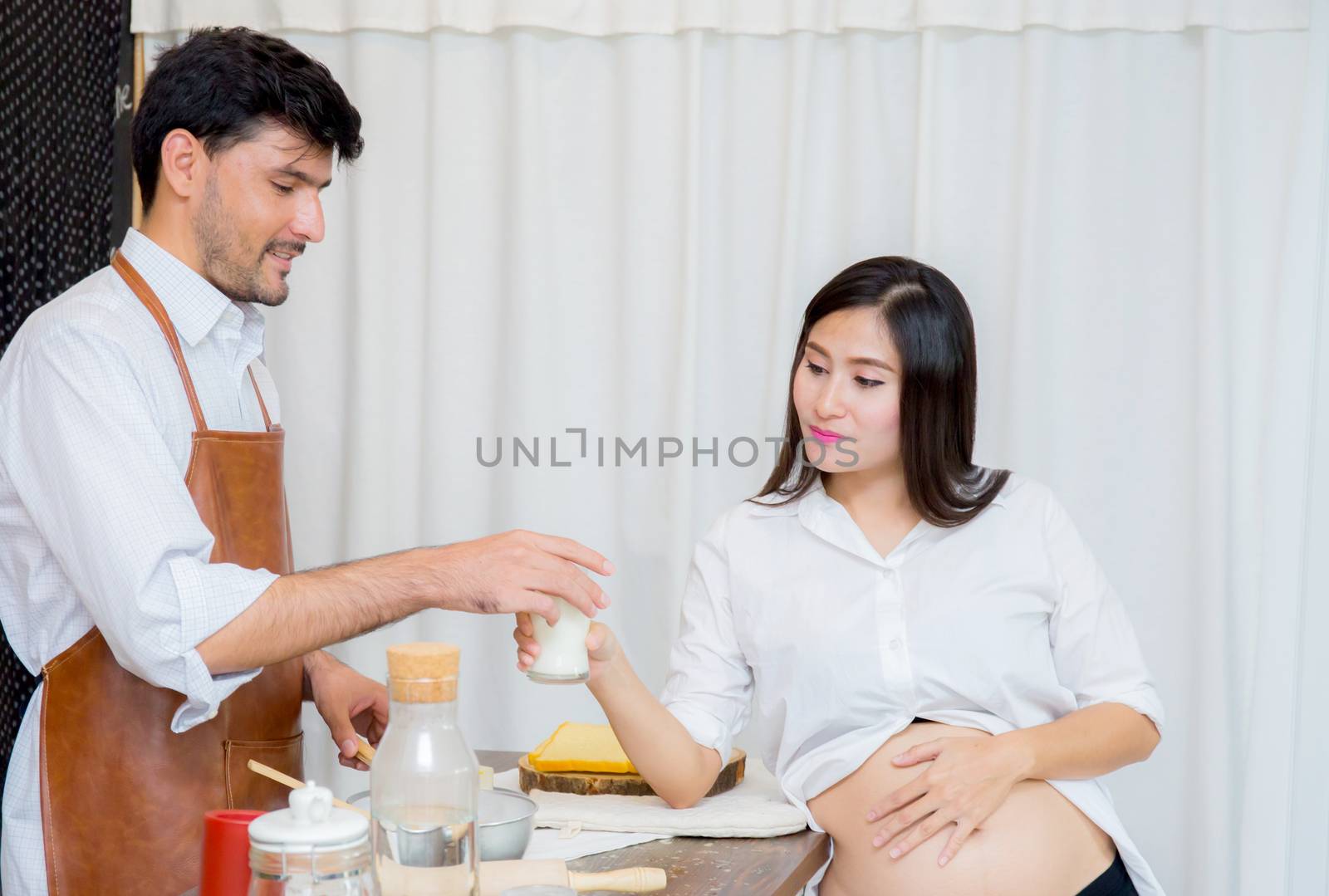 Happy husband and pregnant wife in kitchen,Asian man cooking food in kitchen and woman with drink milk at home,Happy family concept.