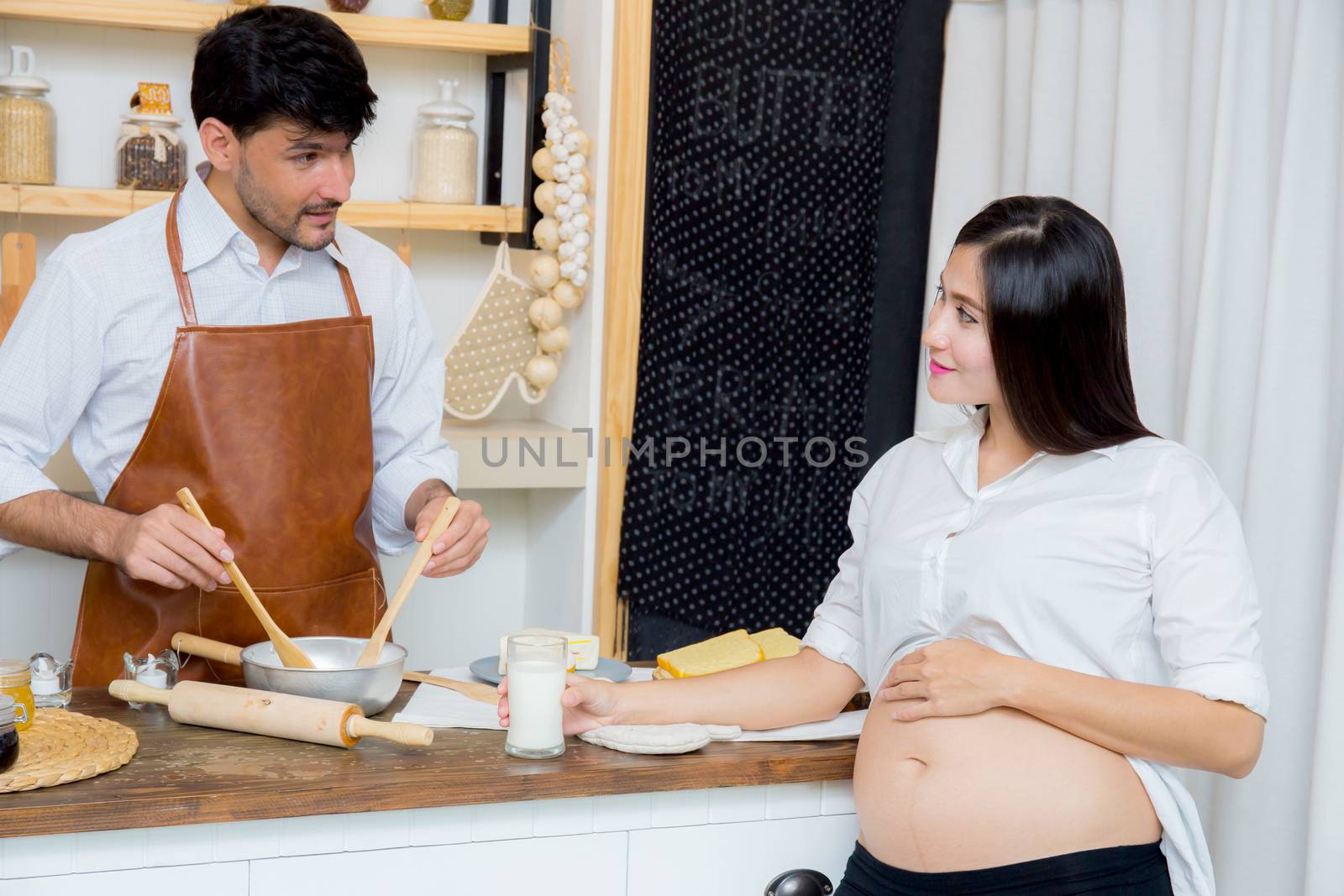 Happy husband and pregnant wife in kitchen,Asian man cooking food in kitchen and woman with drink milk at home,Happy family concept.