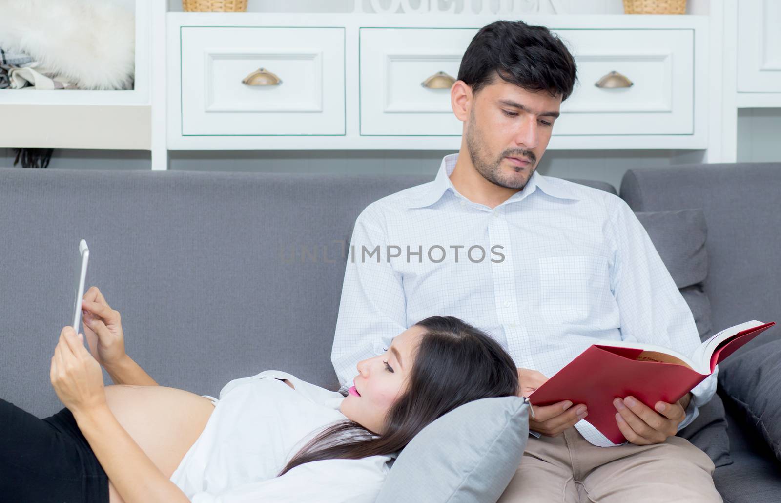 Asian couple family using a tablet at home, the young mother is pregnant lying on sofa looking tablet and husband reading book.