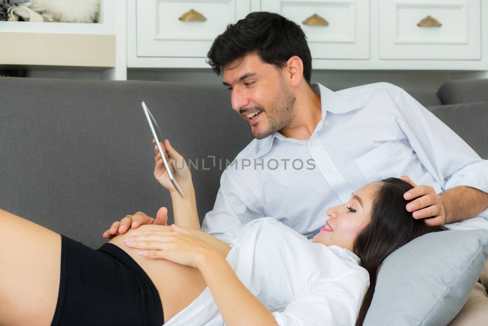Asian couple family young mother and husband using a tablet at home, woman and man is pregnant lying on sofa looking tablet read book.