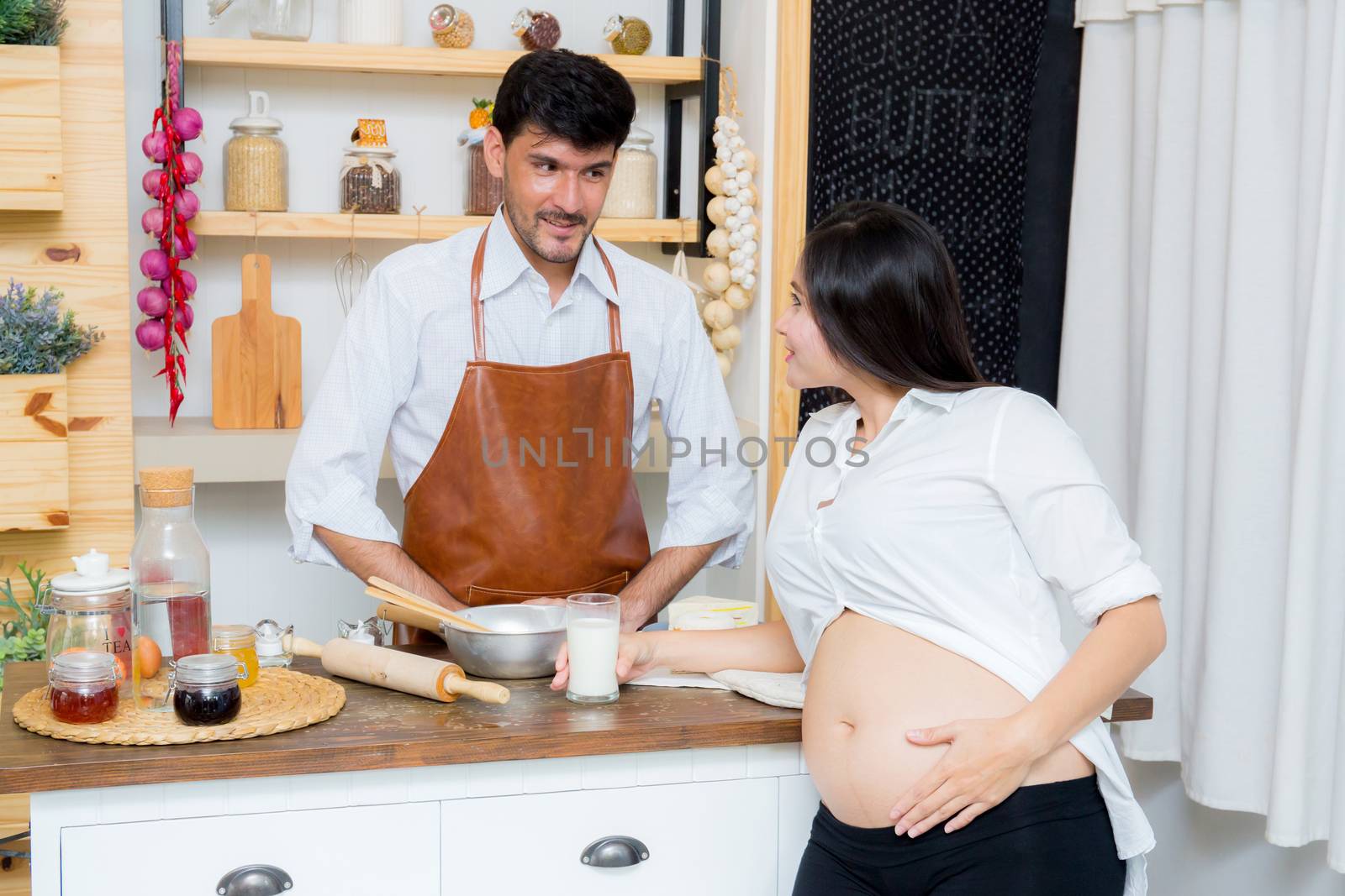 Happy husband and pregnant wife in kitchen,Asian man cooking food in kitchen and woman with drink milk at home,Happy family concept.