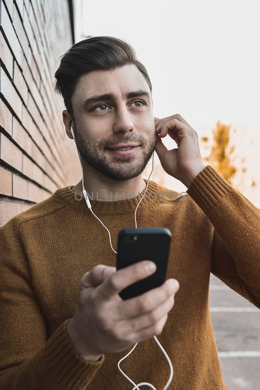 Smiling man listening to music on headphones and leaning against brick wall.