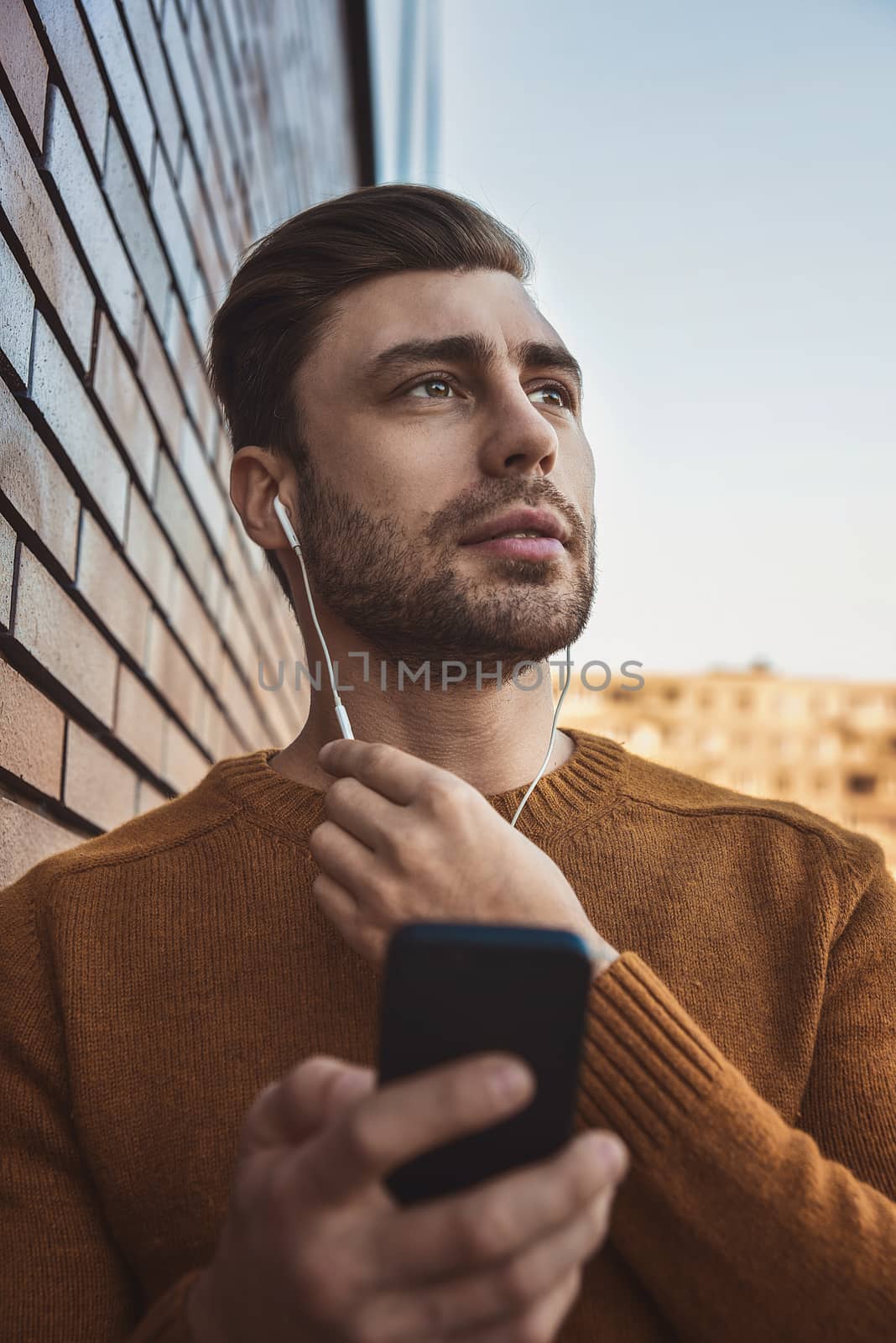 Smiling man listening to music on headphones and leaning against brick wall.