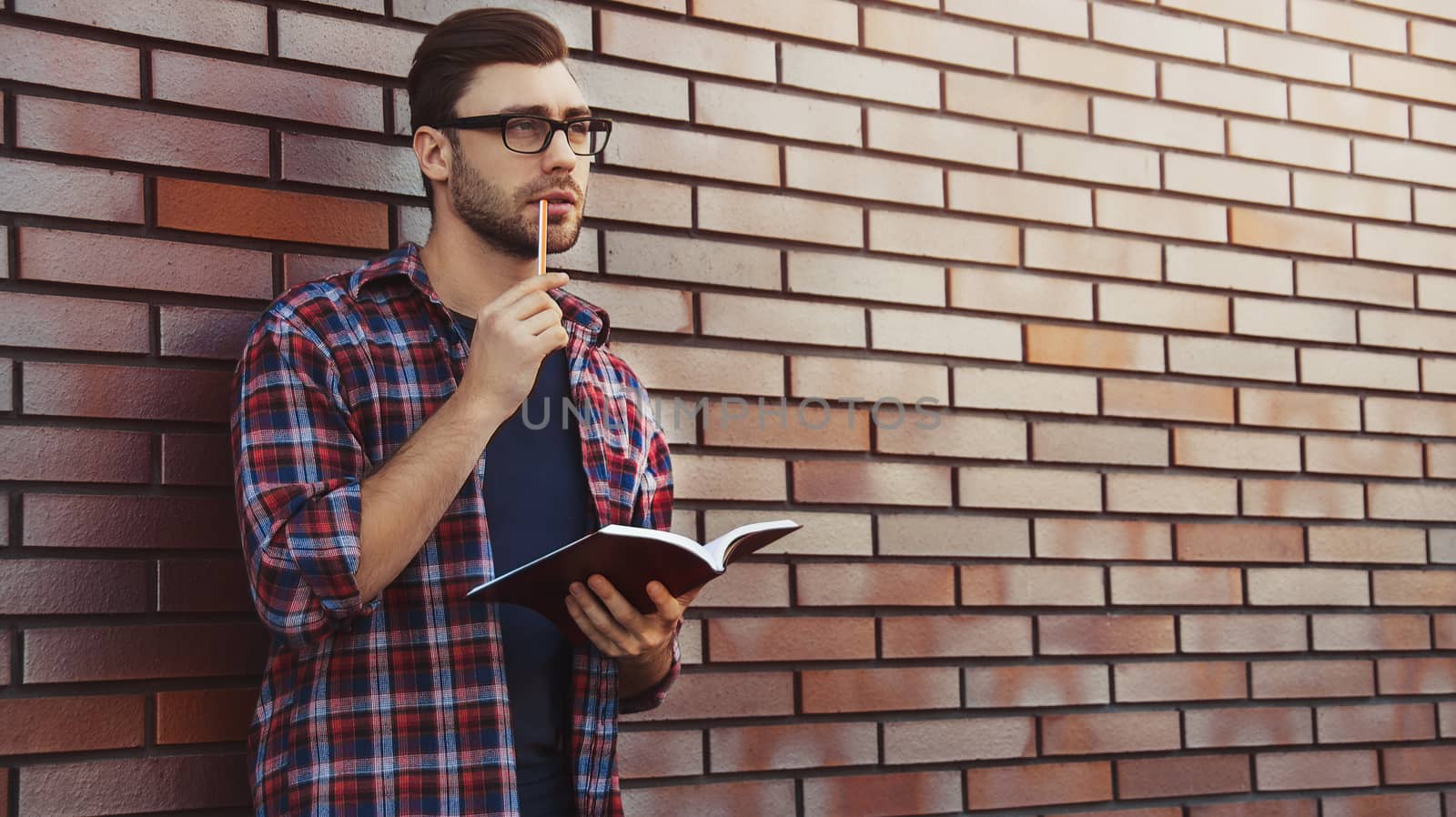 Young hipster guy wearing eyeglasses reading a book or note book make some notes and ideas on brick wall background.