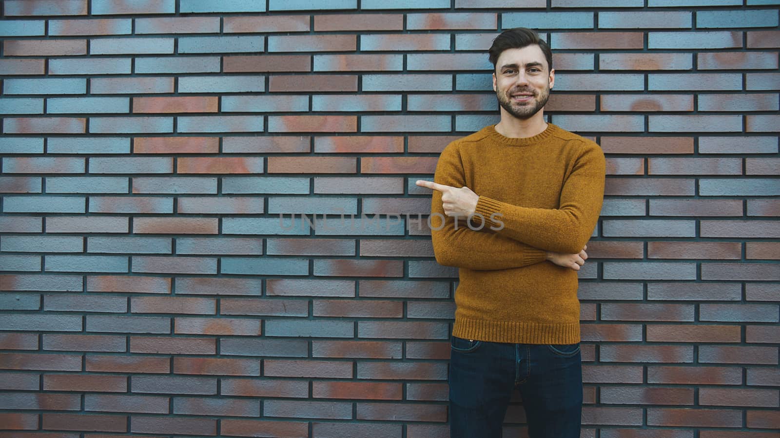 The horizontal look of an attractive young man who advertises something new, points his finger to the side, stands against a brick wall. People and lifestyle