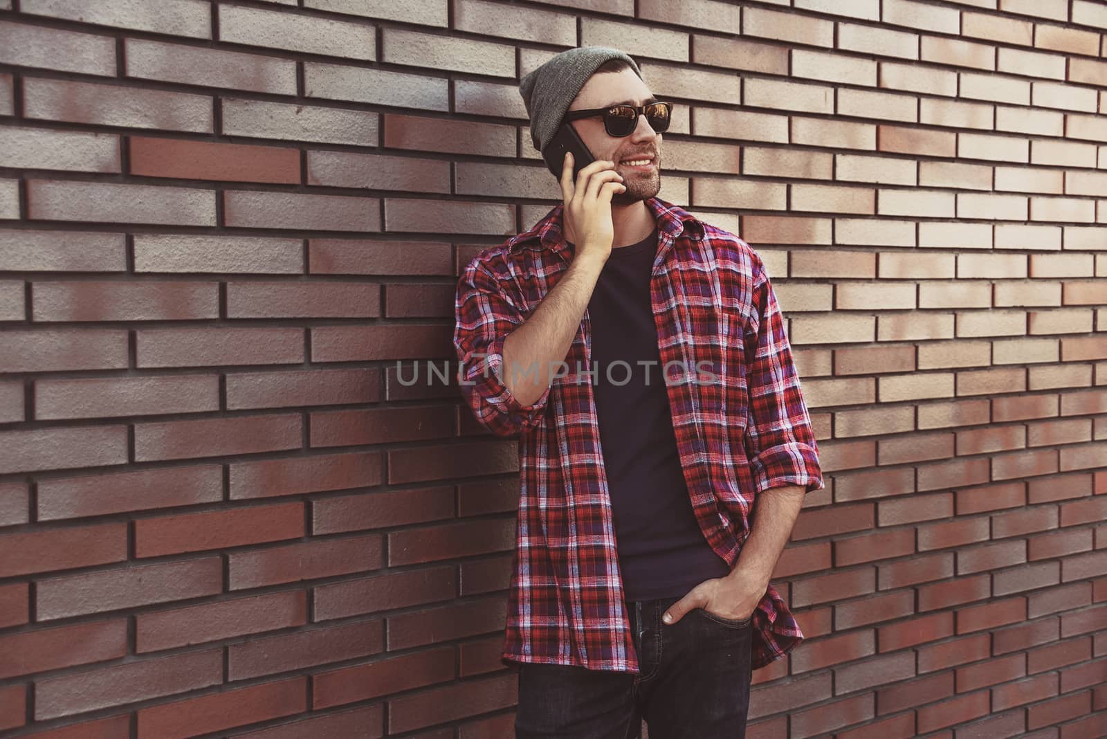 Hipster calling in city street on brick wall background. Amazing man holding smartphone in smart casual wear standing. Urban young professional lifestyle.