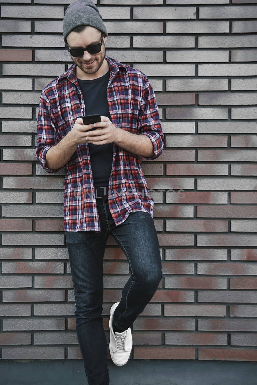 Hipster sms texting phone app in city street on brick wall background. Amazing man holding smartphone in smart casual wear standing.