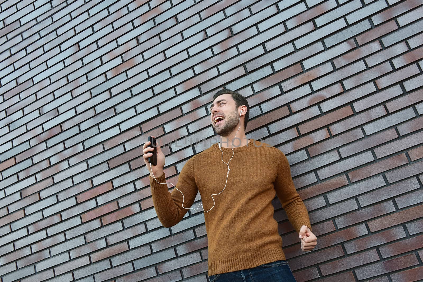 Portrait of smiling man with headphones and cellphone standing by brick wall.