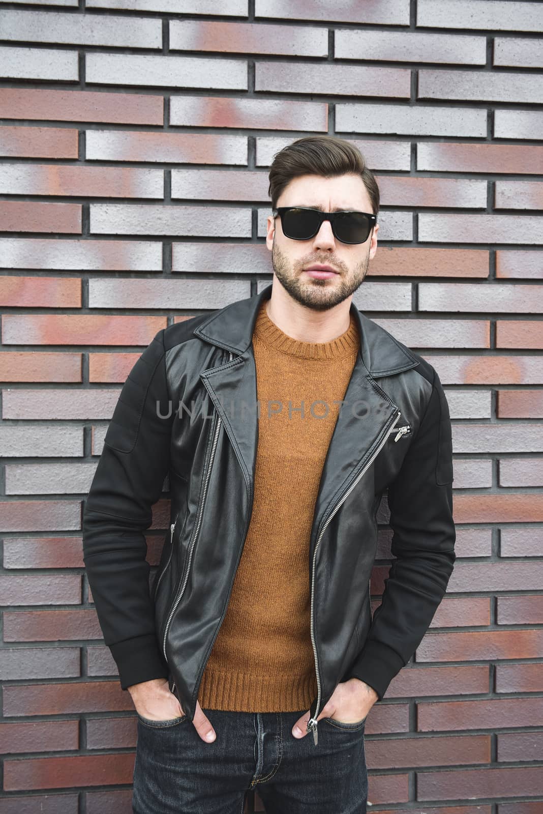 Portrait of stylish handsome young man with bristle standing outdoors and leaning on brick wall.