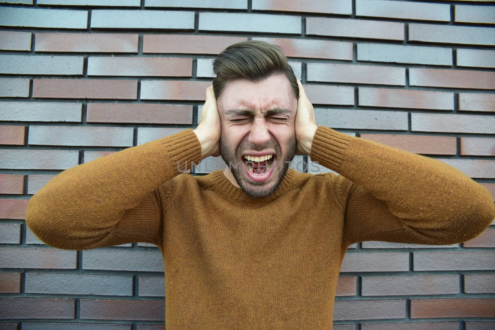 Man feeling painful emotions, screaming, witnessing death of close friend, being miserable and devastated, holding palms on head, yelling from pain and stress, standing unhappy over brown brick wall.
