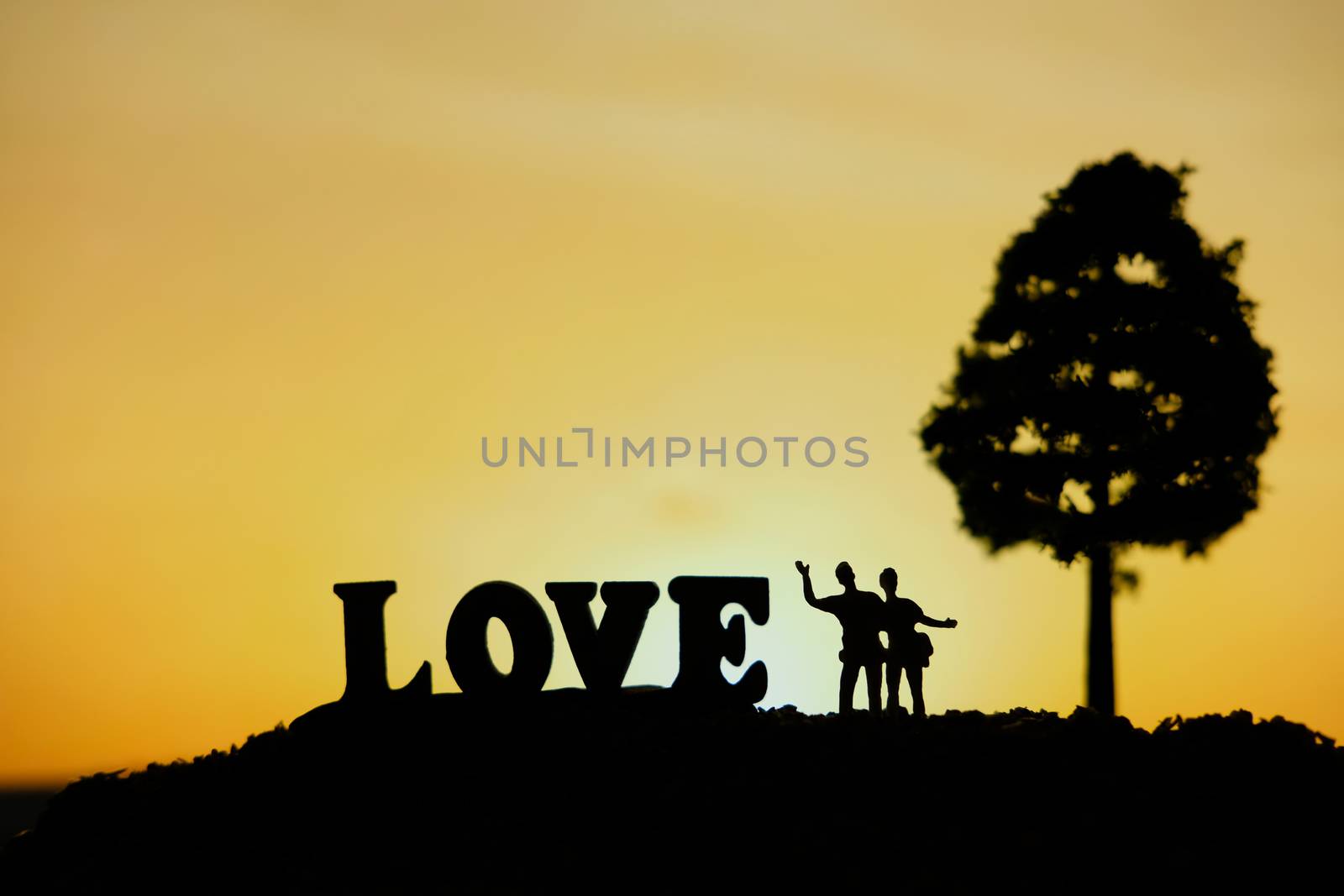 miniature people / toy photography - conceptual valentine holiday illustration. Happy couple holding each other enjoying sunset view under a big tree with love word block