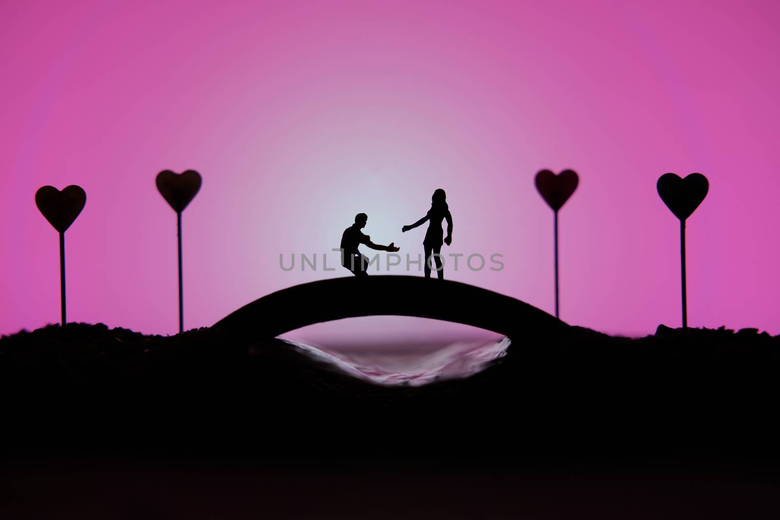 miniature people / toy photography - conceptual valentine holiday illustration. A man proposing a girl silhouette above the bridge with heart lamp by Macrostud