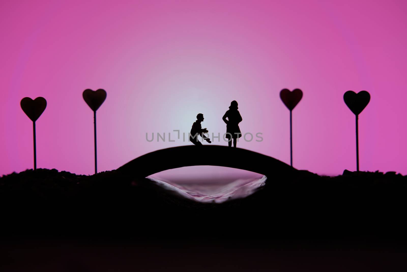 miniature people / toy photography - conceptual valentine holiday illustration. A man proposing a girl silhouette above the bridge with heart lamp