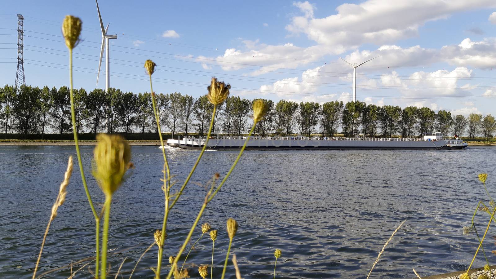 Freight boat water transport passing by on the canal of an inland waterway, surrounded by trees, nature and a wind turbine in the distance. Industry meets nature