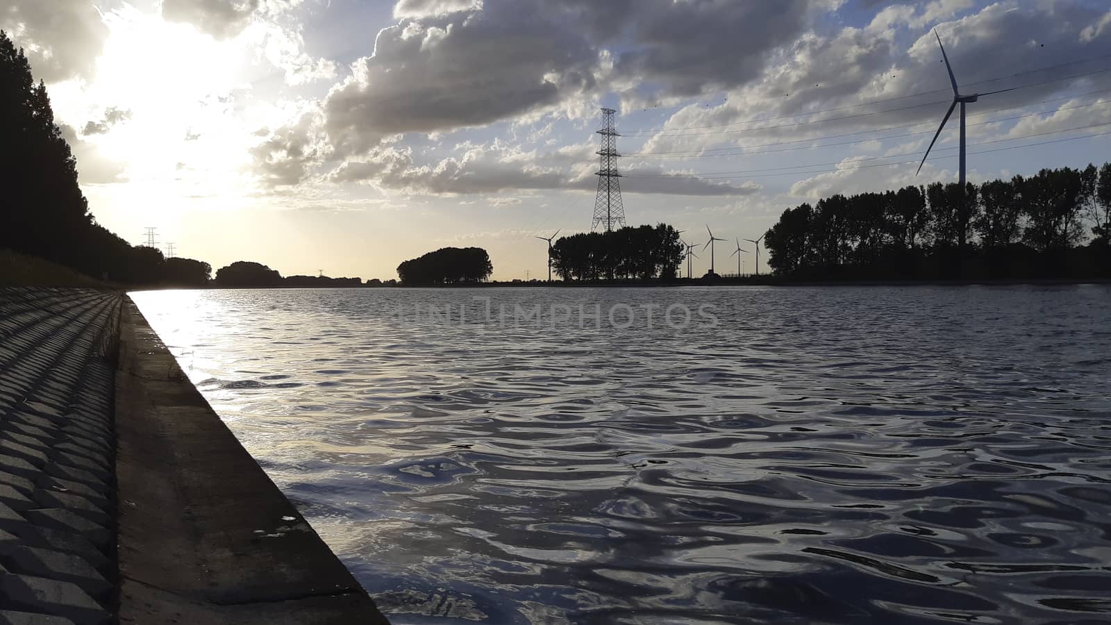 Sunset silhouette on the shore of a canal in industrial area, wind turbine and electricity pylon in the distance.