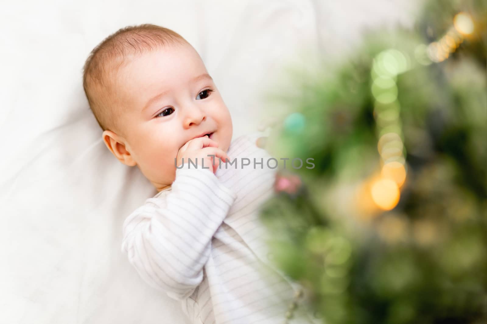Baby boy or girl lying under Christmas tree. Little child plays with fir tree decorated for New Year celebration. Cute kid portrait. Winter holiday spirit.