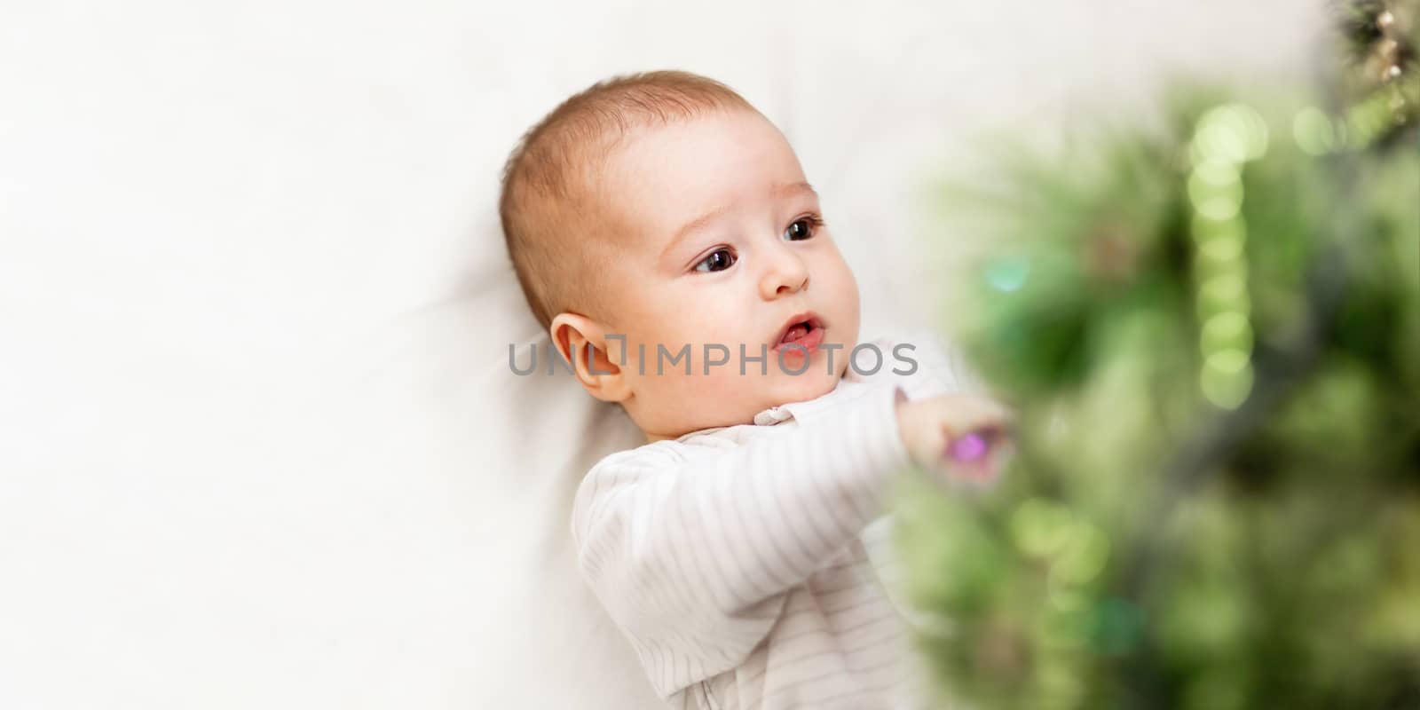 Baby boy or girl lying under Christmas tree. Little child plays with fir tree decorated for New Year celebration. Cute kid portrait. Winter holiday spirit. Banner with copy space.
