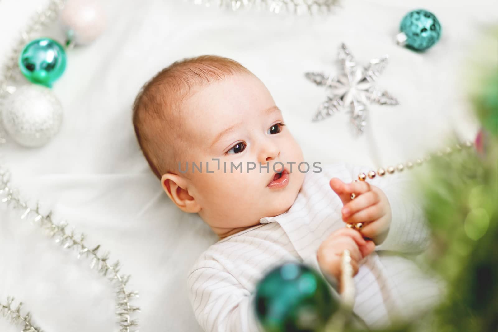 Baby boy or girl lying under Christmas tree. Little child plays with fir tree decorated for New Year celebration. Cute kid portrait. Winter holiday spirit.