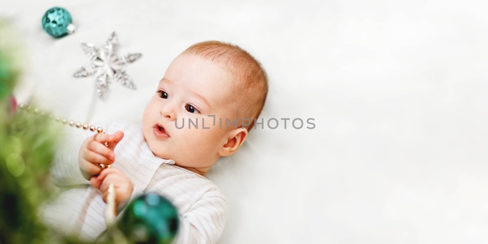 Baby boy or girl lying under Christmas tree. Little child plays with fir tree decorated for New Year celebration. Cute kid portrait. Winter holiday spirit. Banner with copy space.