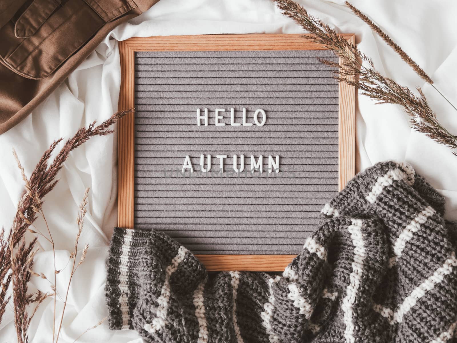 Top view on letter board with season greeting HELLO AUTUMN. Crumpled textile background with dried grass, grey knitted sweater and brown warm outfits.