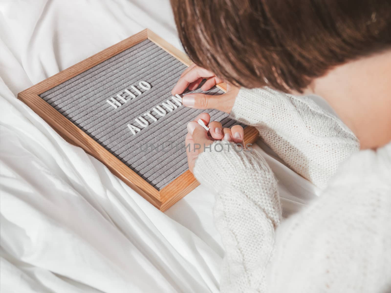 Woman in white knitted sweater typing HELLO AUTUMN on letter board. Crumpled textile background. Season greeting concept. Sunny, warm and cozy home.
