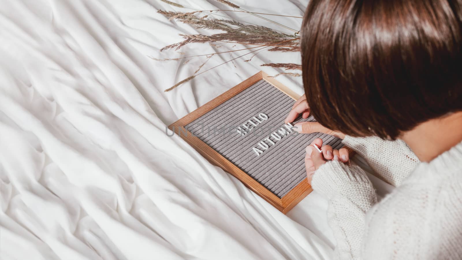 Woman in white knitted sweater typing HELLO AUTUMN on letter board. Crumpled textile background with dried grass. Season greeting banner with copy space. Sunny, warm and cozy home.