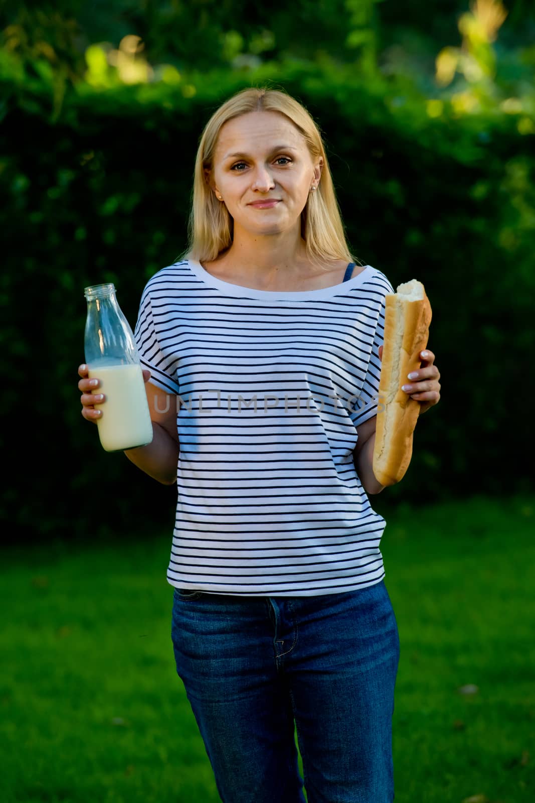 Portrait of a young woman who stands in her garden and holds a glass bottle with milk and a baguette.