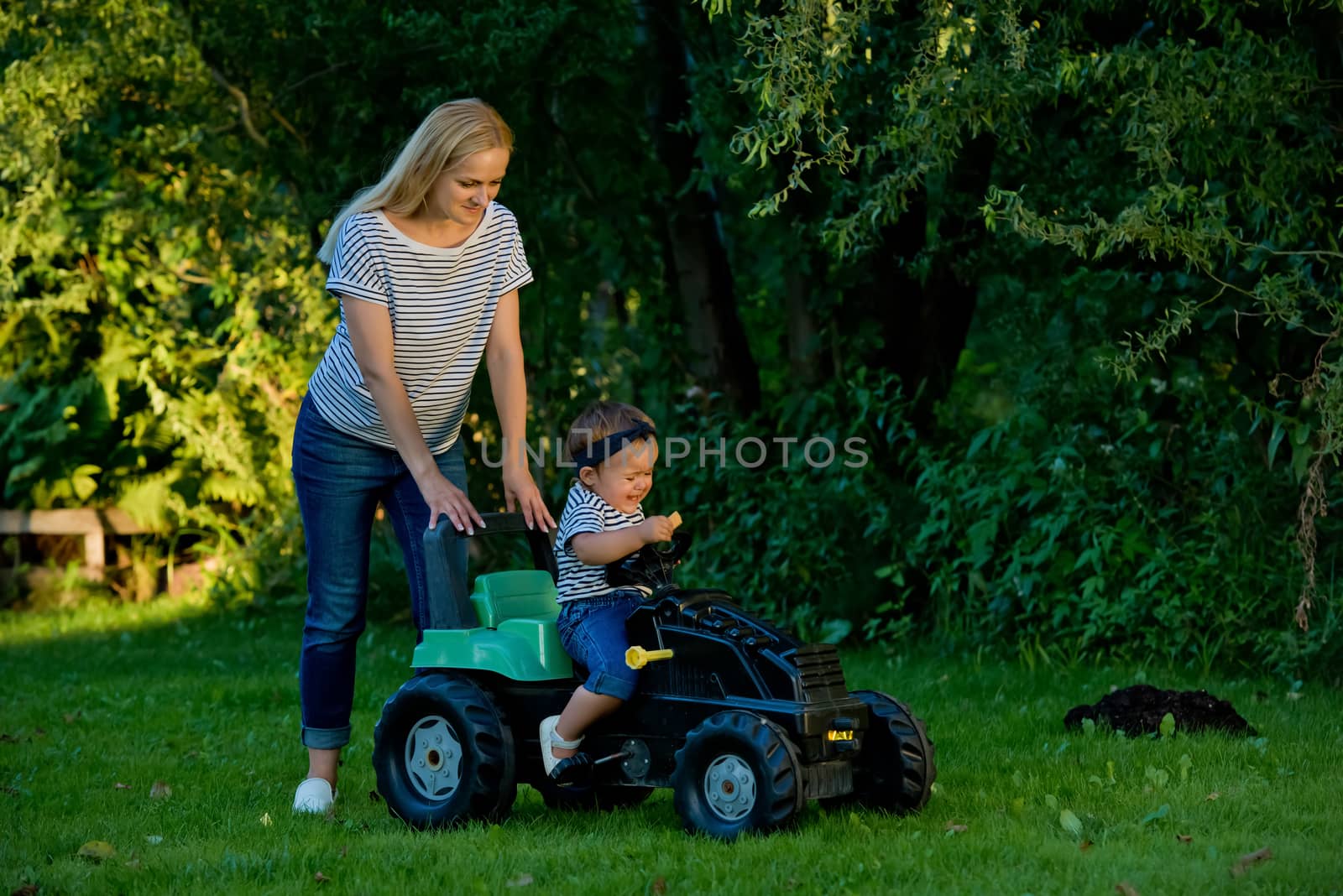 Baby girl and mother playing with toy tractor in a garden.