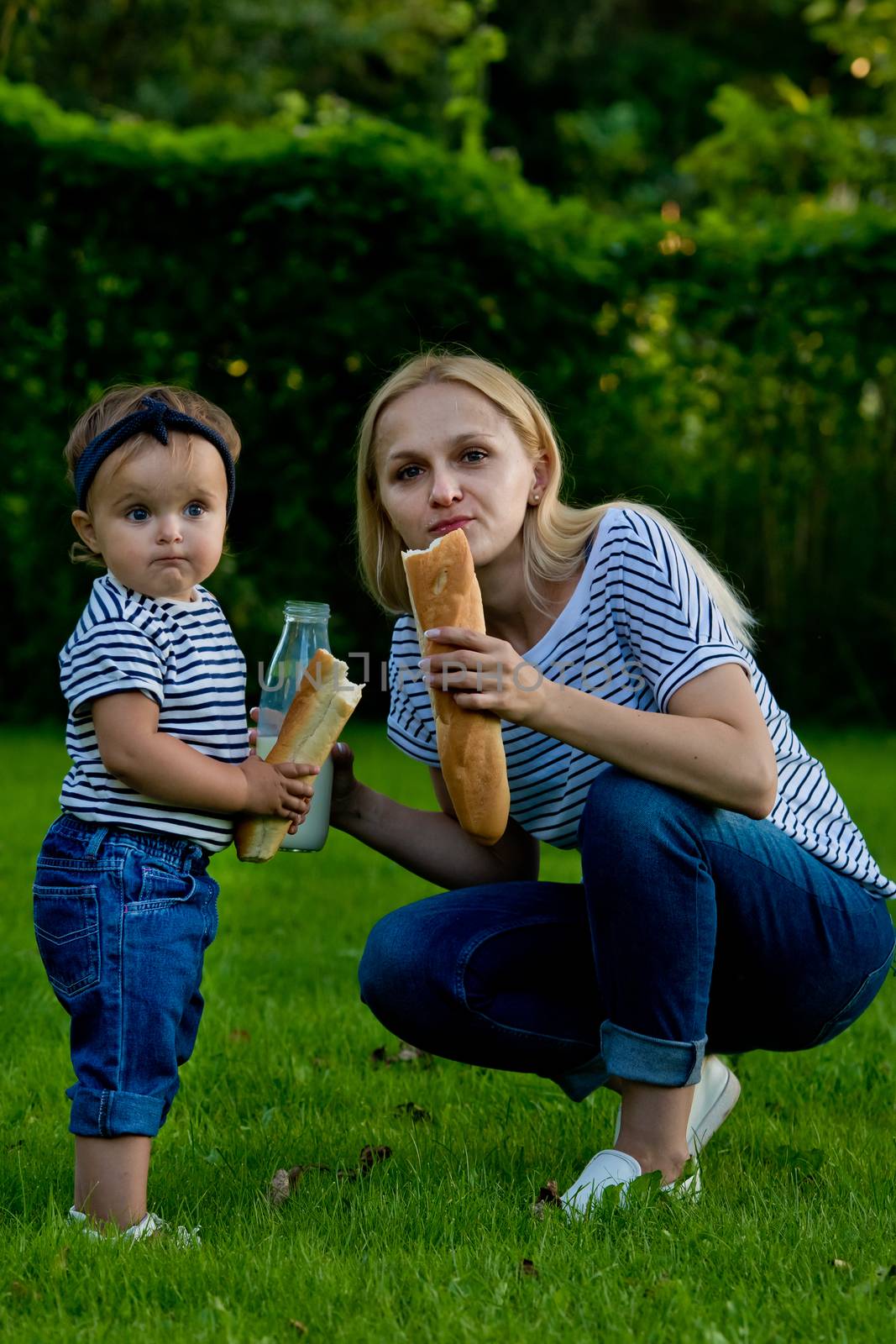 A young woman in jeans and a striped T-shirt gives her daughter milk from a glass bottle. Family picnic. by leonik