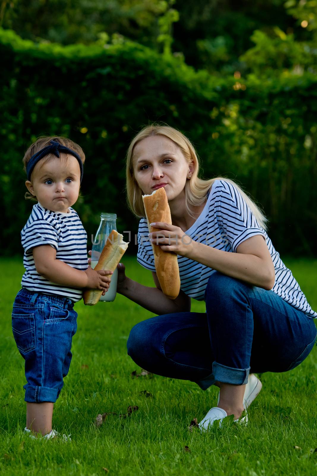 A young woman in jeans and a striped T-shirt gives her daughter milk from a glass bottle. Family picnic.