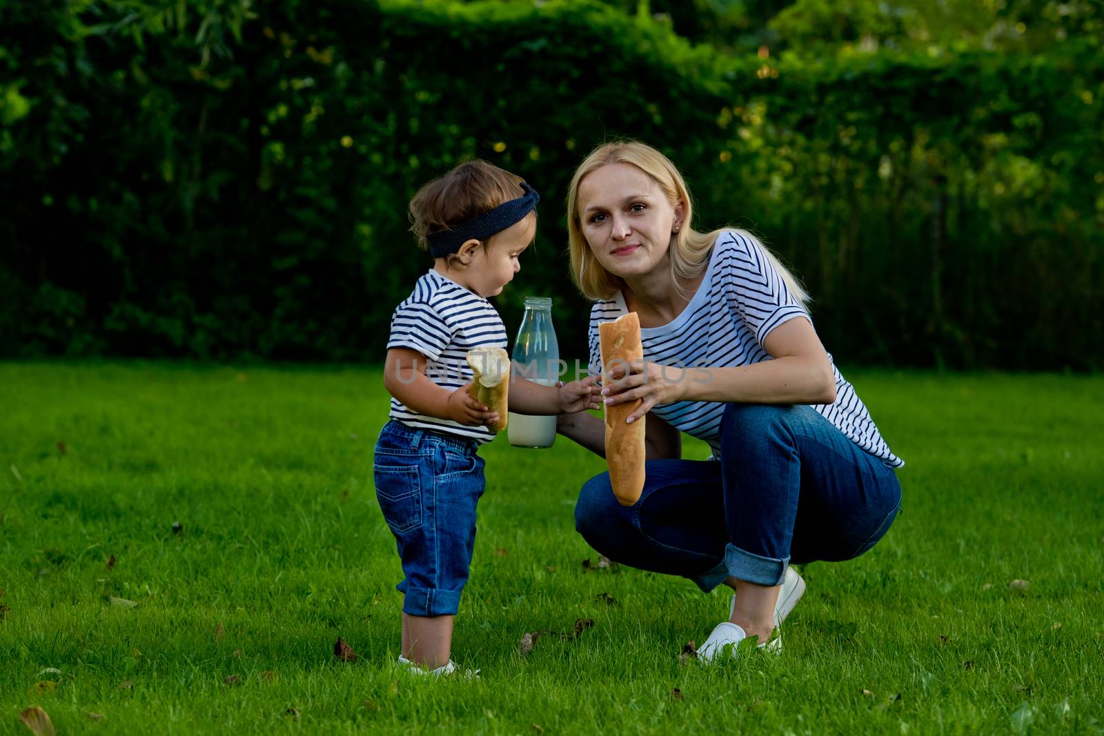 A young woman in jeans and a striped T-shirt gives her daughter milk from a glass bottle. Family picnic.