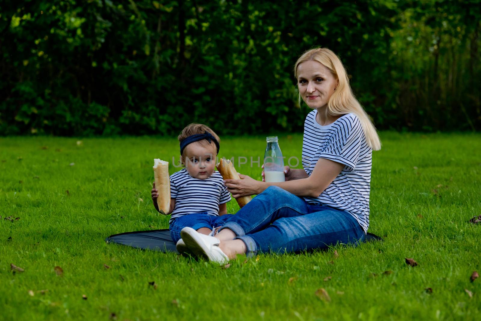 A young woman in jeans and a striped T-shirt gives her daughter a fresh baguette to eat. Family picnic.