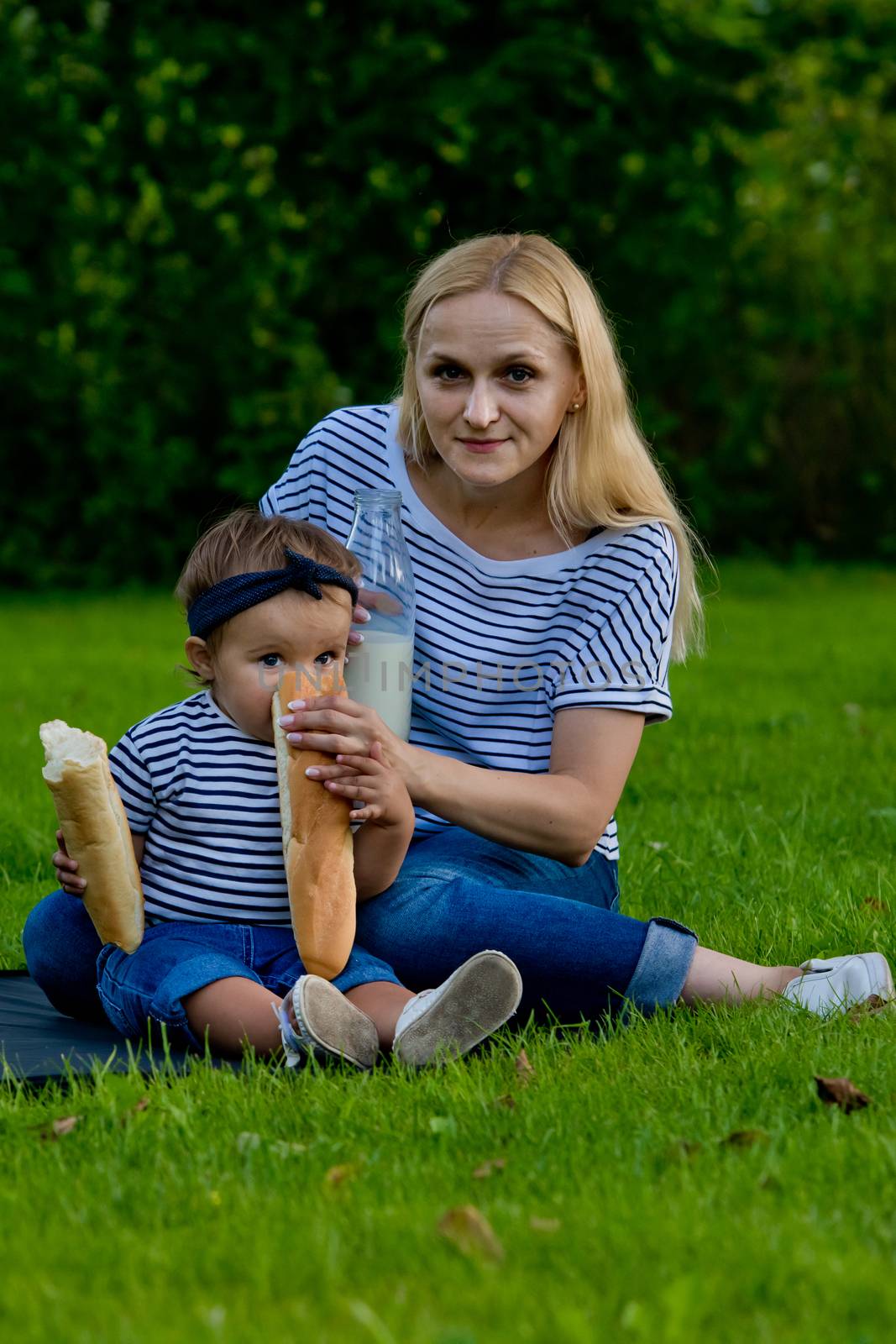 A young woman in jeans and a striped T-shirt gives her daughter a fresh baguette to eat. Family picnic.
