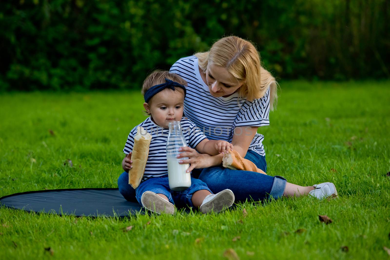 A young woman in jeans and a striped T-shirt gives her daughter milk from a glass bottle. Family picnic.