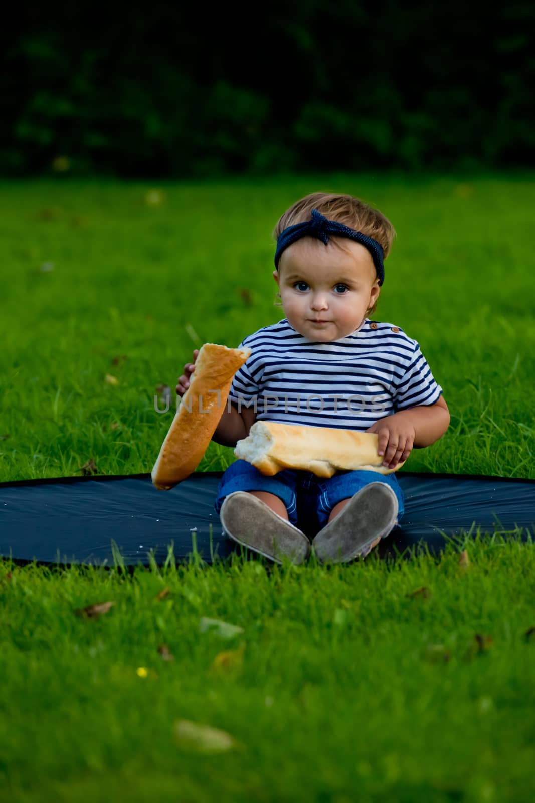 A little pretty baby girl sits on the lawn in the garden and holds a fresh baguette.