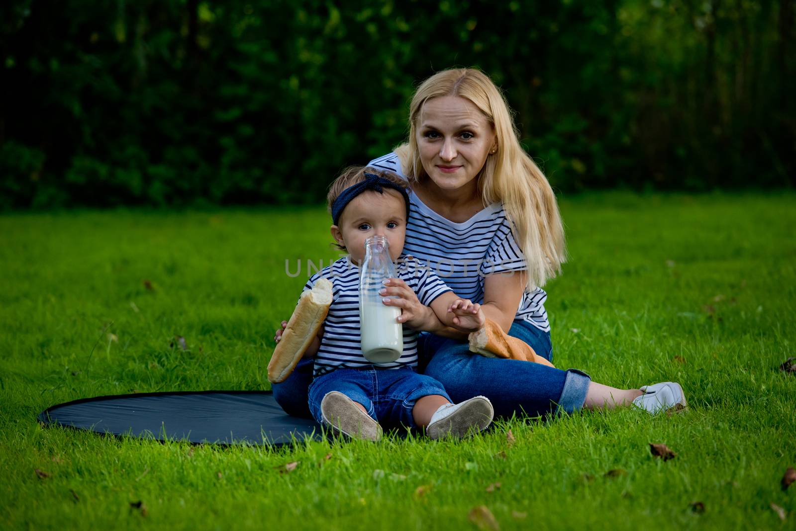 A young woman in jeans and a striped T-shirt gives her daughter milk from a glass bottle. Family picnic.