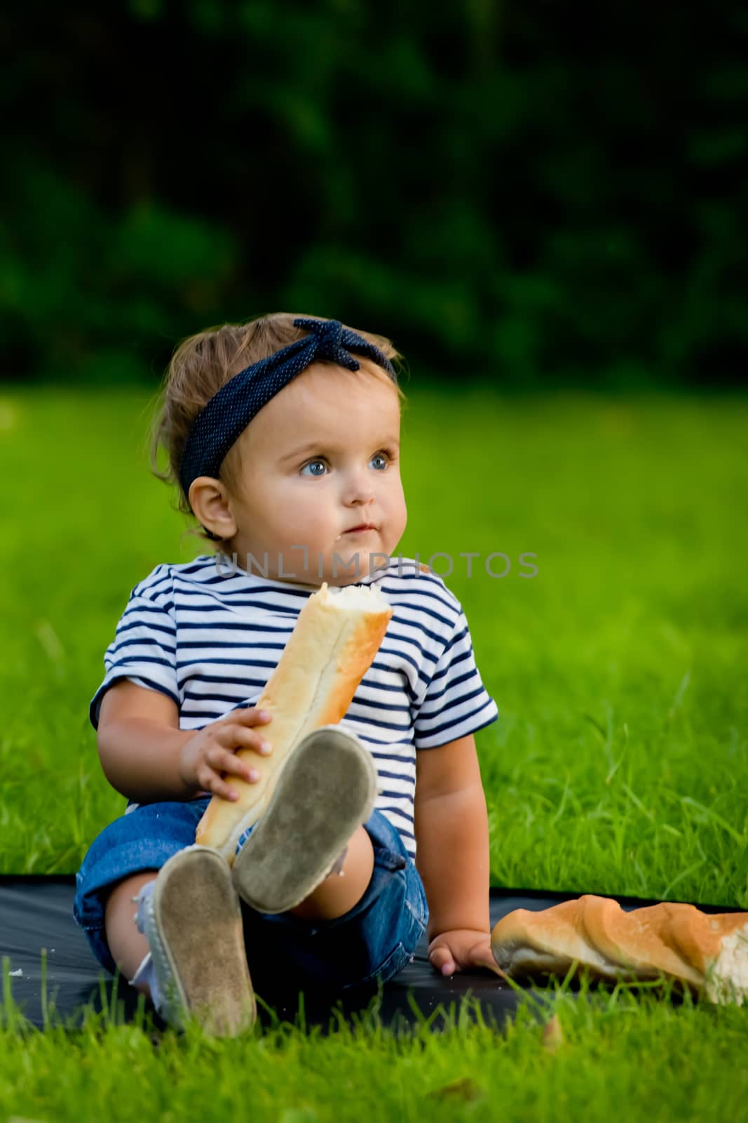 A little pretty baby girl sits on the lawn in the garden and holds a fresh baguette.