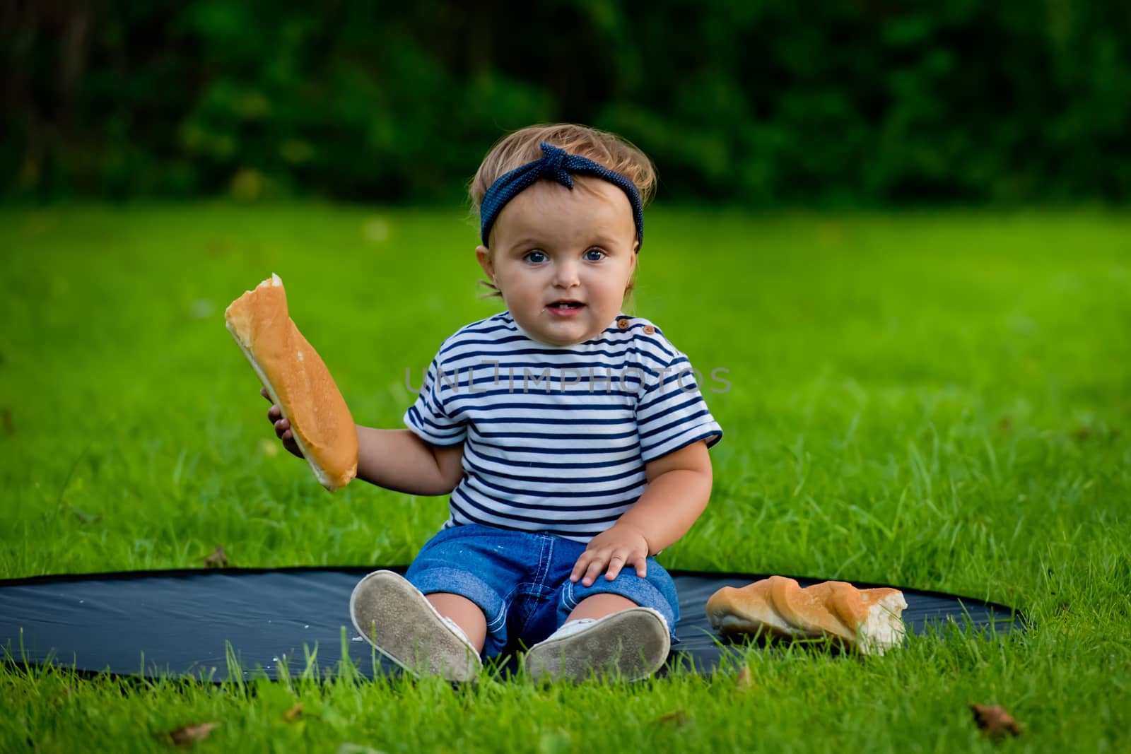 A little pretty baby girl sits on the lawn in the garden and holds a fresh baguette.