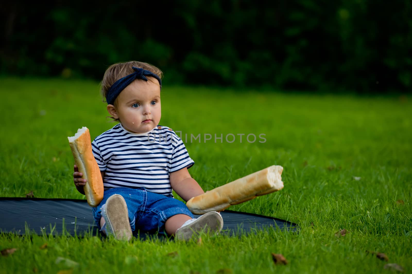 A little pretty baby girl sits on the lawn in the garden and holds a fresh baguette.