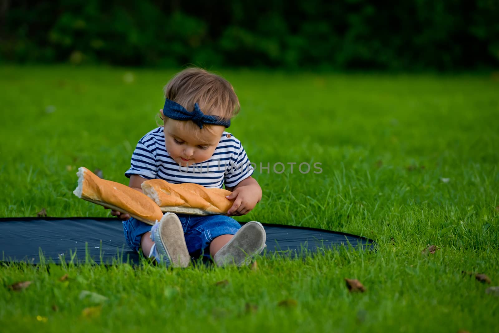 A little pretty baby girl sits on the lawn in the garden and holds a fresh baguette.