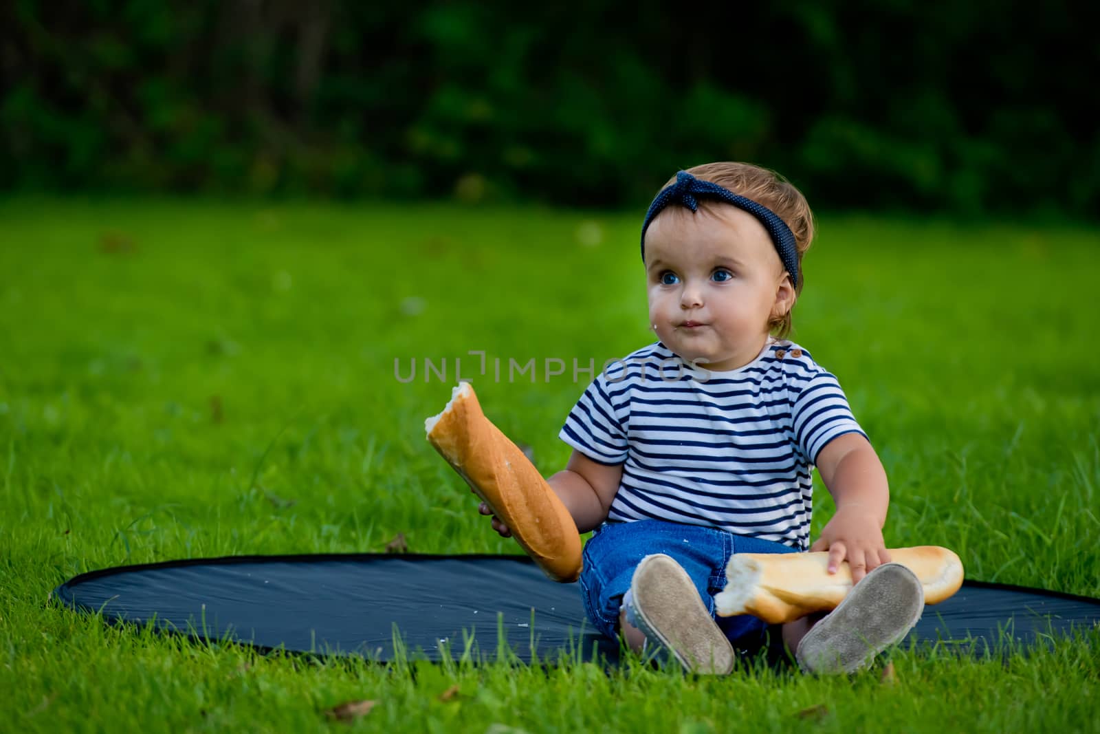 A little pretty baby girl sits on the lawn in the garden and holds a fresh baguette.