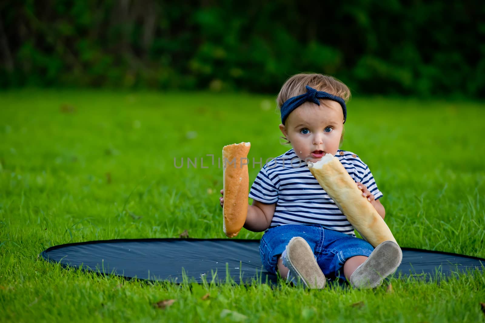 A little pretty baby girl sits on the lawn in the garden and holds a fresh baguette. by leonik