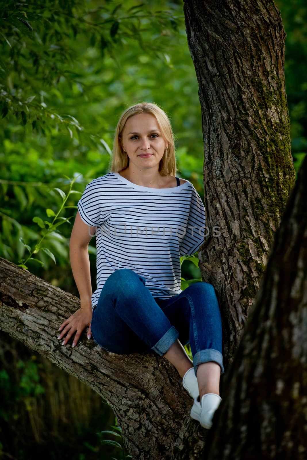 A young woman in jeans and a striped T-shirt sits on a tree.