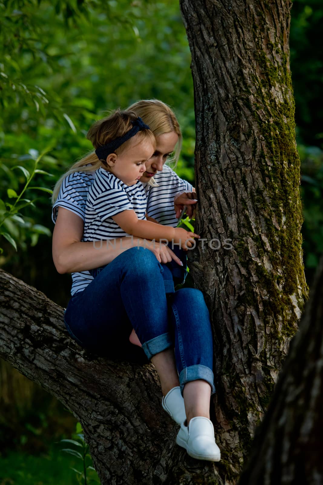 A young woman in jeans and a striped T-shirt and her little daughter are sitting on a tree.