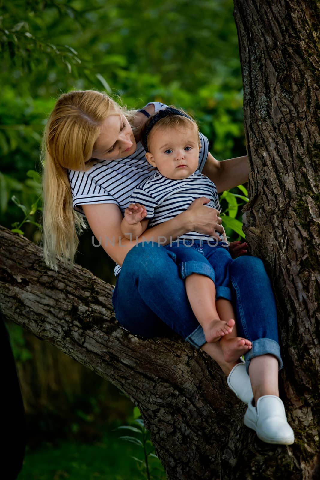 A young woman in jeans and a striped T-shirt and her little daughter are sitting on a tree.