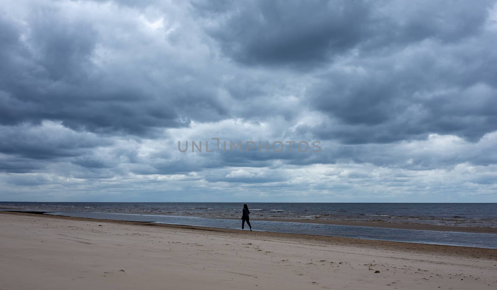 A girl in a warm black jacket walks along the seashore on a sandy beach in cloudy, cool weather.